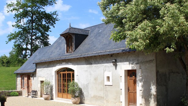 A rustic stone building with a sloped roof, large wooden doors, and a gravel driveway.