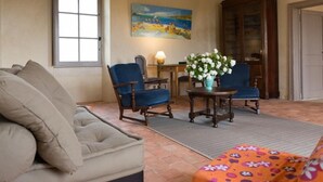 Una sala de estar acogedora con un sofá color beige, un sillón azul y un reposapiés estampado con motivos florales. Se observa un cuadro en la pared, un aparador de madera y una lámpara sobre una mesita.