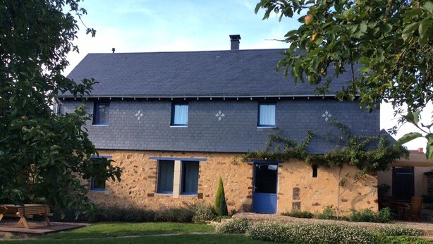 Une maison à deux étages, dotée d’un toit en pente, d’une façade en pierre et d’un petit jardin avec un banc.