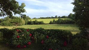 Un jardin bordé d’une haie de fleurs rouges, avec vue sur un champ verdoyant où sont entreposées des bottes de foin.