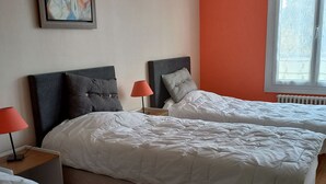 A hotel room with two beds, each with a grey headboard and white bedding. There is a red lamp on a wooden nightstand beside each bed. A framed picture hangs on the wall above the beds. - Spacieuse maison confortable avec jardin, à 20 min du centre-ville et proche du circuit des 24H du Mans (Le Mans)