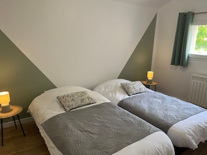 A room with two beds, each with a patterned pillow, a bedside table with a lamp, and a window with curtains. - Cottage in the countryside near Le Mans (Brette-les-Pins)