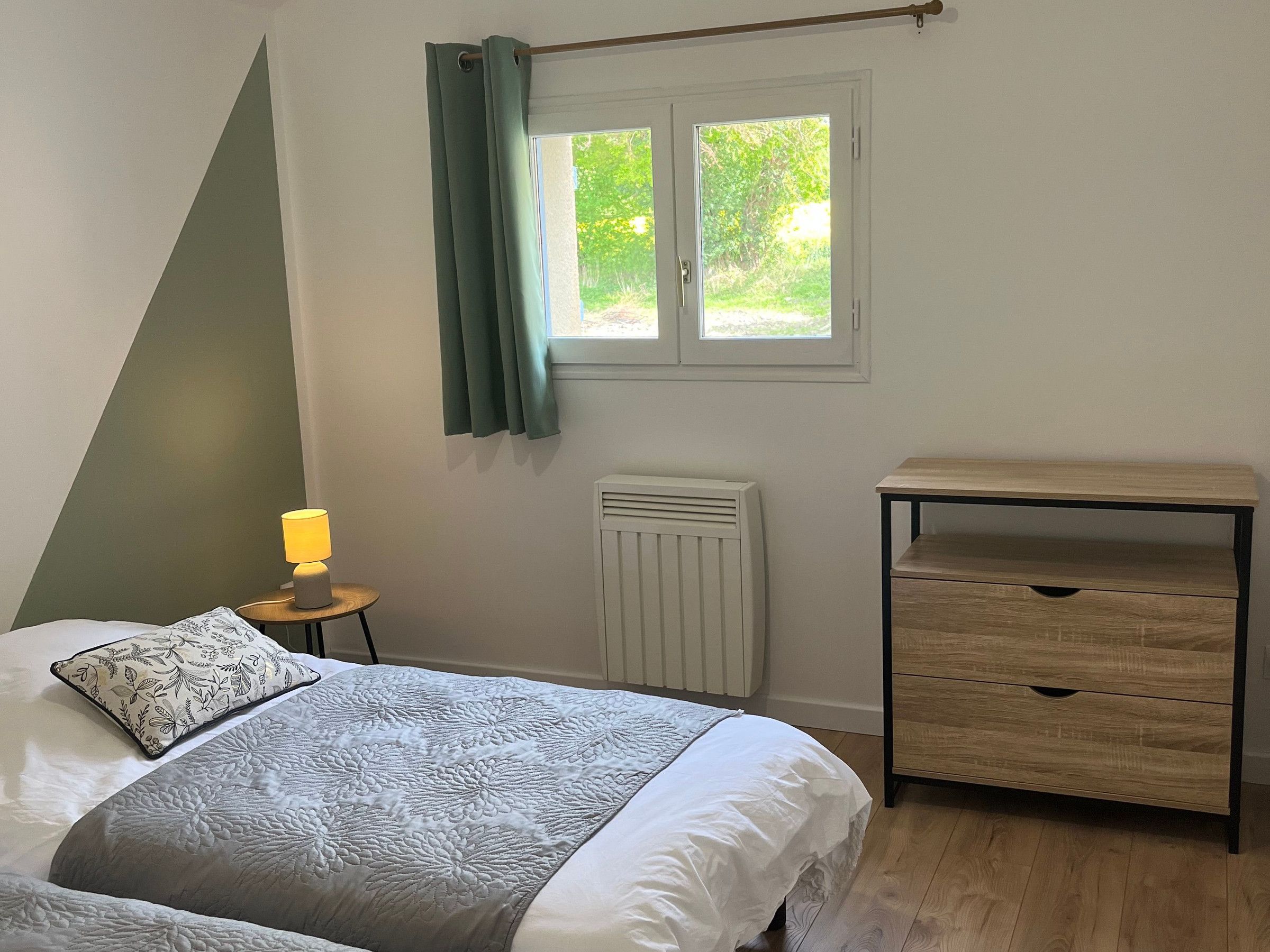 Une chambre à coucher comprenant un lit, une table de chevet avec une lampe, un radiateur, une fenêtre avec des rideaux et une commode en bois.
