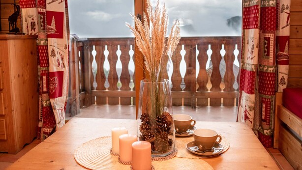 Ein Holztisch mit Kerzen, einer Vase mit getrockneten Pflanzen und zwei Tassen auf einem Balkon mit gemusterten Vorhängen.
