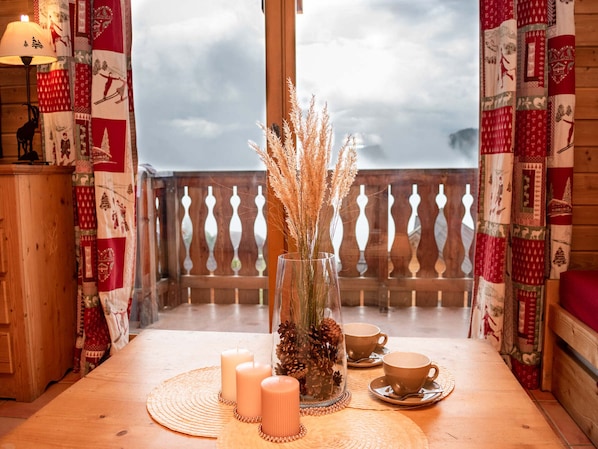 A wooden table with candles, a vase with dried plants, and two cups on a balcony with patterned curtains.