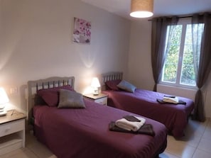 Een slaapkamer met twee bedden, elk met een nachtkastje en een lamp. Er is een raam met gordijnen en een aan de muur bevestigd kunstwerk.