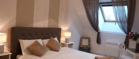 A neatly made bed with pillows, a bedside table with a lamp, a chair, and a window with curtains.