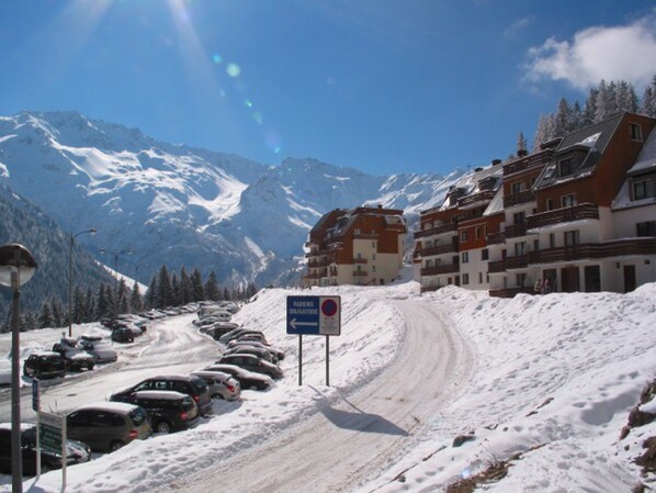 A snow-covered road with parked cars, a sign indicating parking is obligatory, and buildings with a mountainous backdrop.