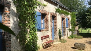 A rustic building with brick walls and blue shutters, a bench, and a garden area with a fence and sheep.