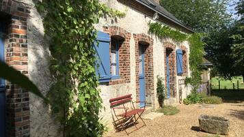 Een landelijk gebouw met bakstenen muren en blauwe luiken, een bankje en een tuin met een hek en schapen.