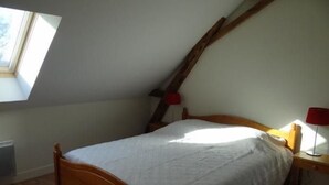 Quarto com teto inclinado, cama de madeira, mesa de cabeceira com abajur e janela que ilumina naturalmente.