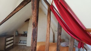 Un dormitorio tipo loft con techo inclinado, vigas de madera, una cama con ropa de cama naranja y blanca, un aparador de madera y un dosel rojo.