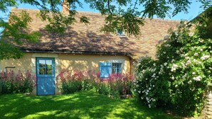 Una casa rústica con techo de tejas, un pequeño porche y un jardín con flores rosas y blancas.