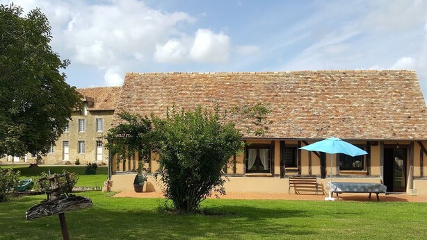Un bâtiment avec un toit recouvert de tuiles, une pelouse verdoyante, un banc et un parasol bleu.
