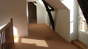 Un couloir étroit, recouvert de moquette, avec des rampes en bois et un plafond en pente, qui mène à une pièce dotée d’une fenêtre.