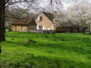 Un bâtiment rustique en pierre, doté d’un toit en pente, entouré d’une pelouse verdoyante et luxuriante et d’arbres en fleurs.