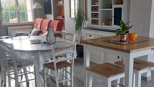 Uma área de refeições com uma mesa redonda, cadeiras brancas, uma bancada de cozinha pequena e vista para o exterior pela janela.