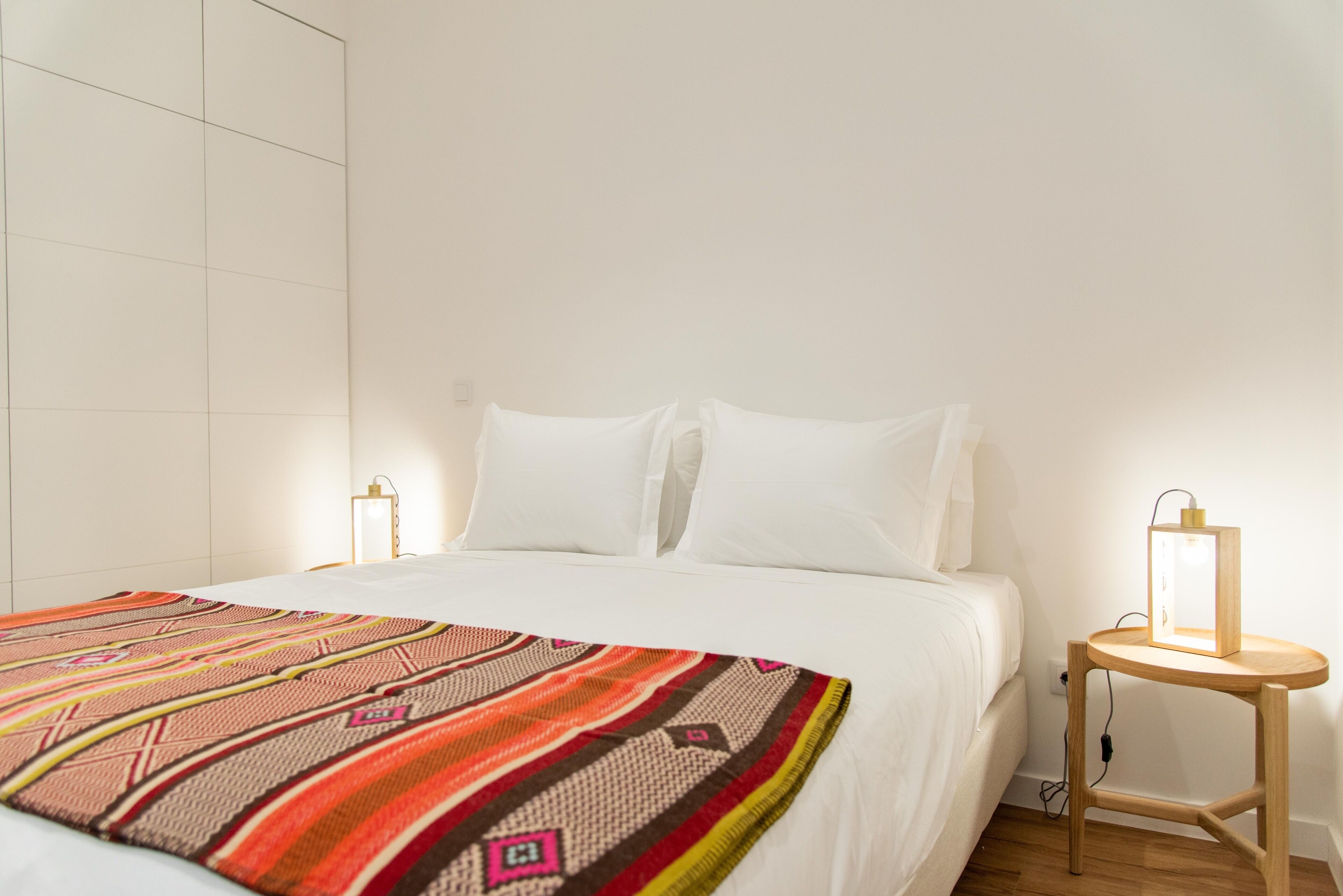 A neatly made bed with a colorful blanket, a bedside table with a lamp, and a wall-mounted light fixture.