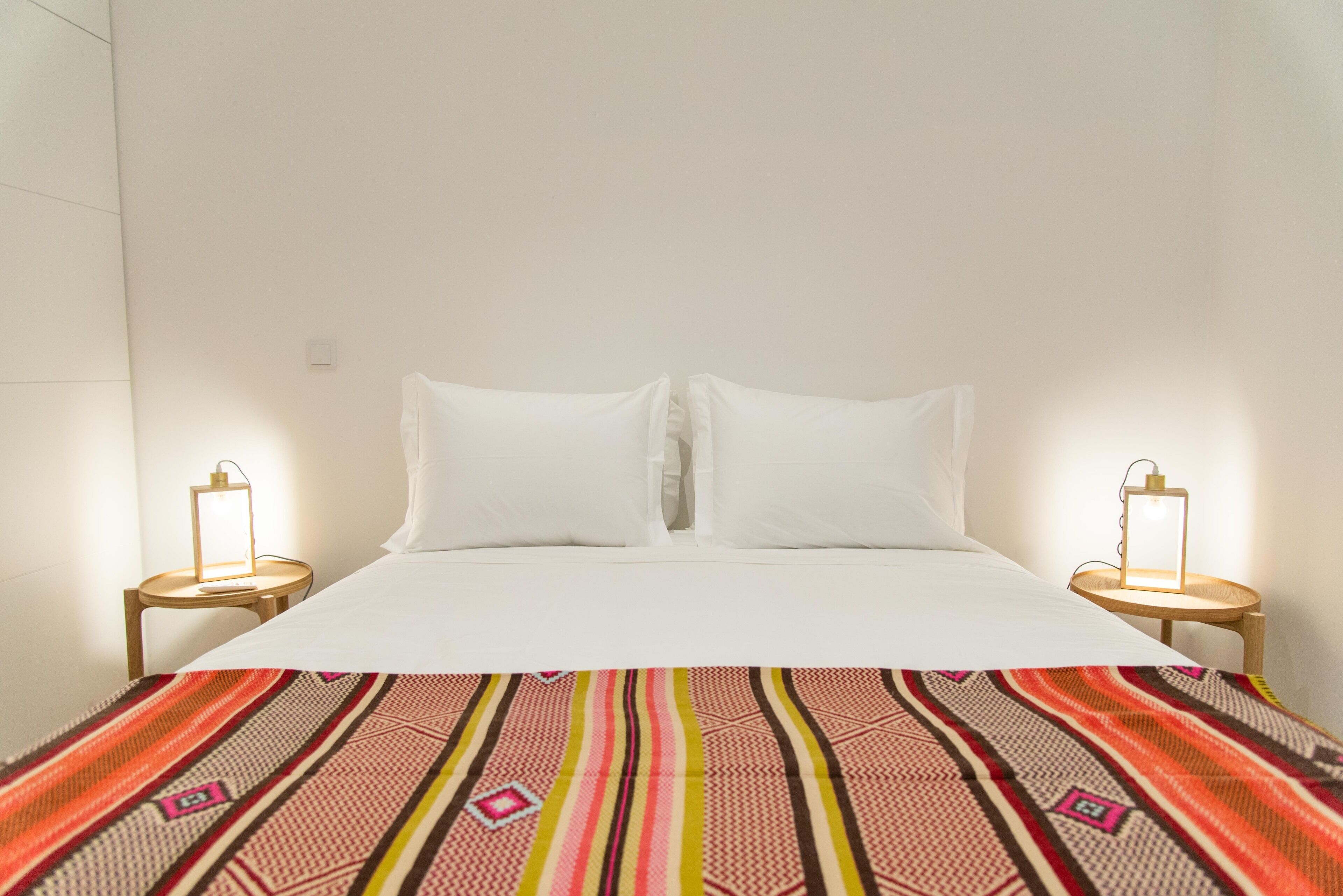 A bed with white linens and a colorful striped blanket, two bedside lamps, and a plain white wall.