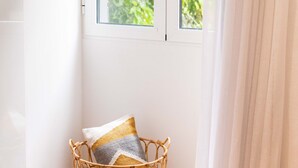 A corner room with a white wall, a wicker chair with a cushion, and a window with white curtains. - Apartamentos Fernão Ornelas (Funchal)