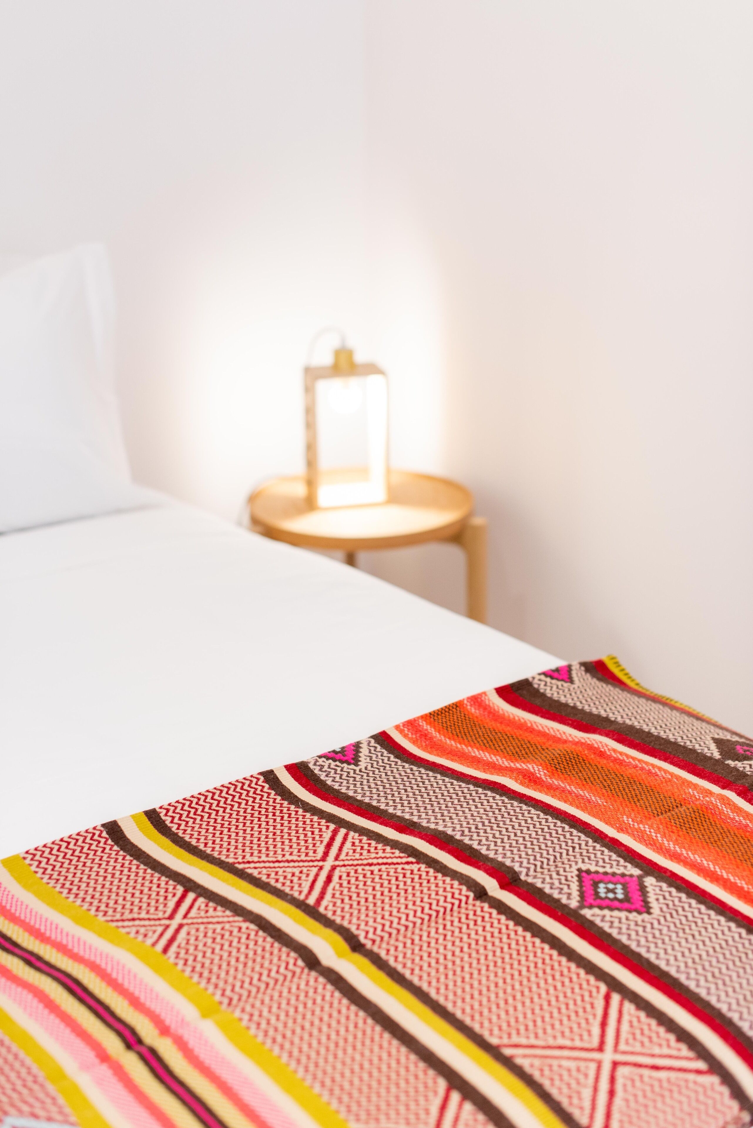 Ein ordentlich bezogenes Bett mit einer farbenfrohen Decke, ein Nachttisch mit einer Lampe und eine weiße Wand.