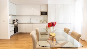 A modern kitchen with white cabinets, a microwave, and a dining area with a glass table and beige chairs. - Apartamentos Fernão Ornelas (Funchal)