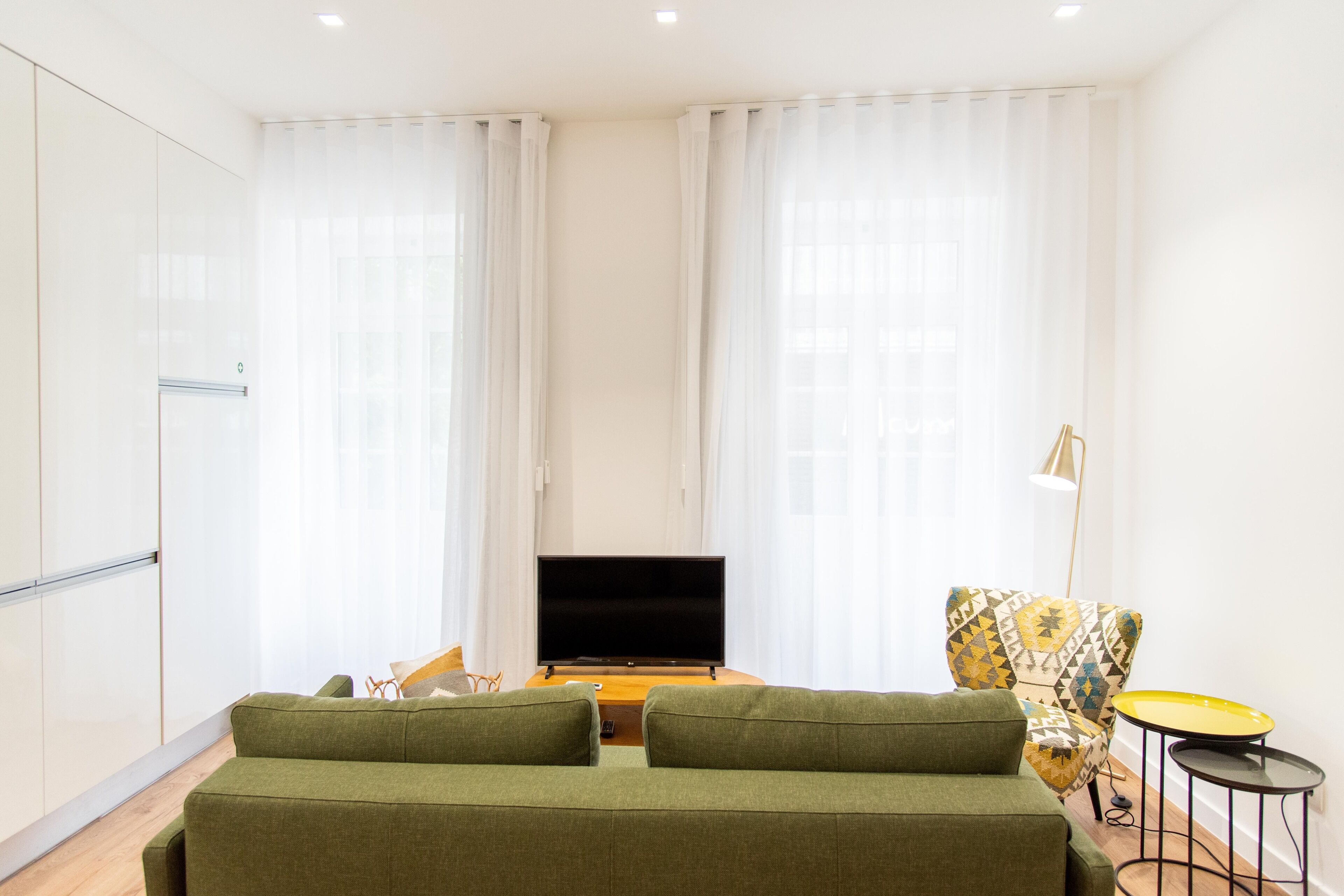 A modern hotel room with a green sofa, a flat-screen TV, a patterned armchair, and a small round table.
