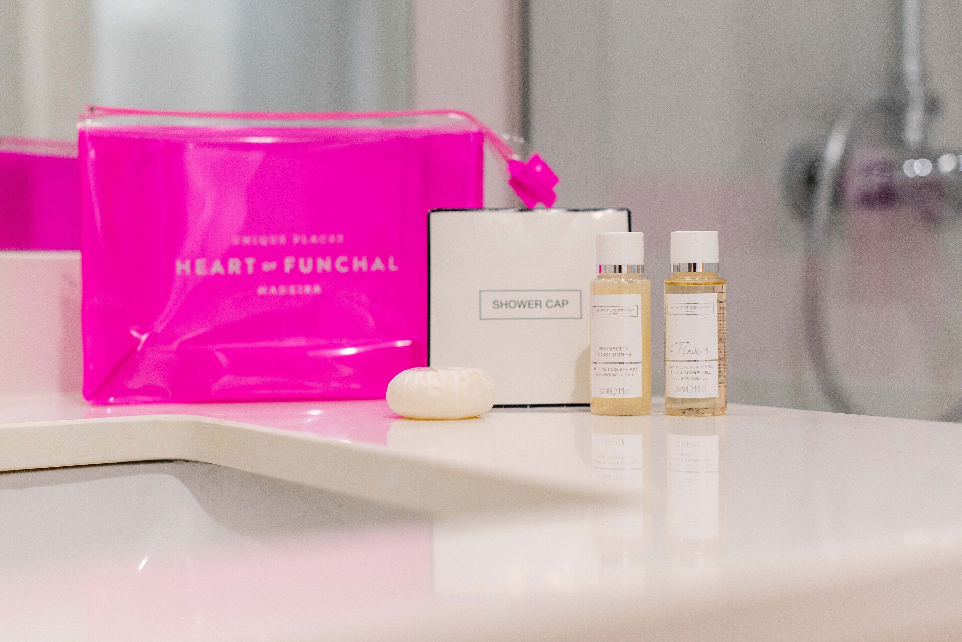 A bathroom counter with a pink bag labeled "Heart of Funchalf," a shower cap, and two bottles of shampoo and conditioner.