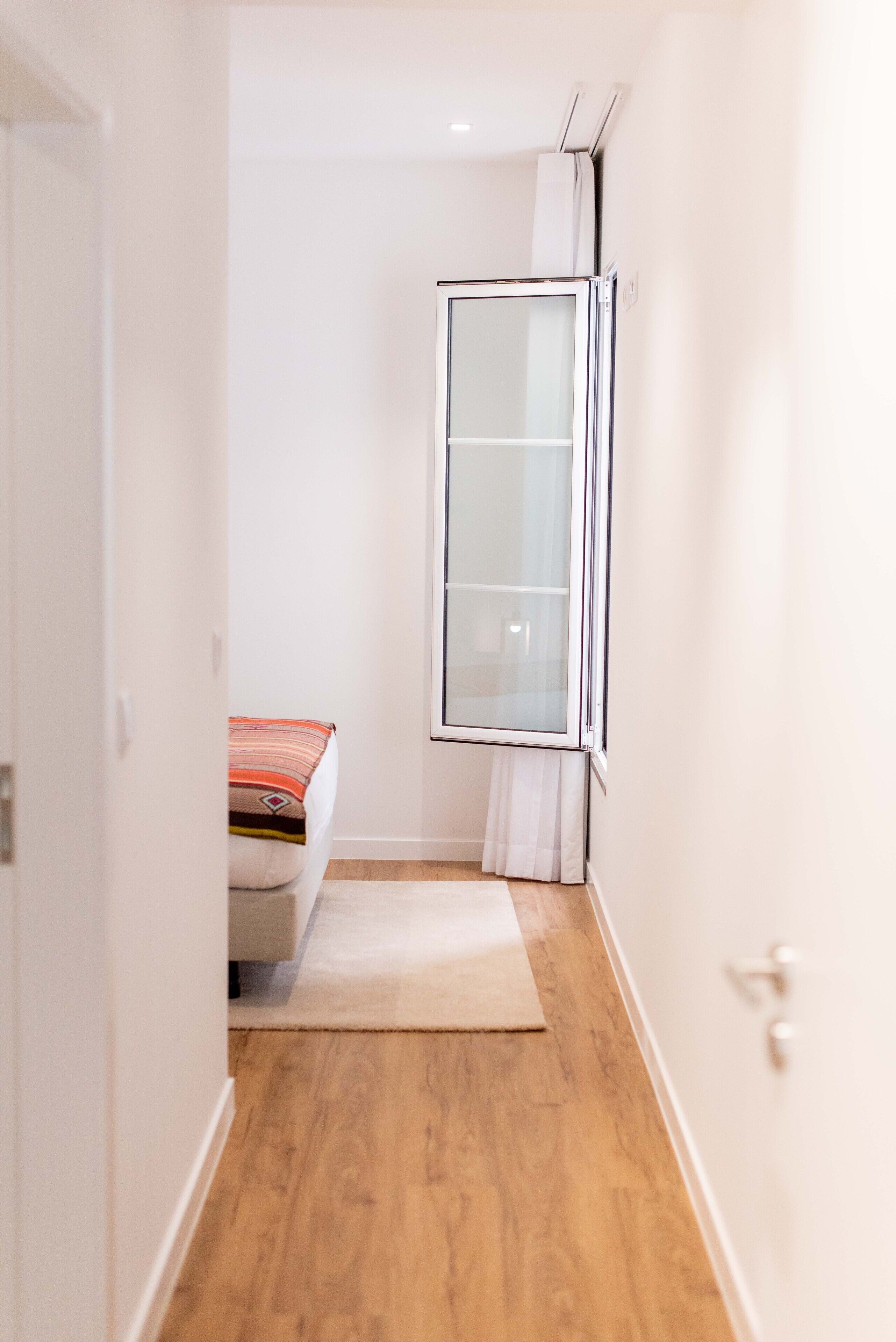 Un pasillo estrecho con suelo de madera, una cama con manta a rayas y una ventana con persiana blanca.