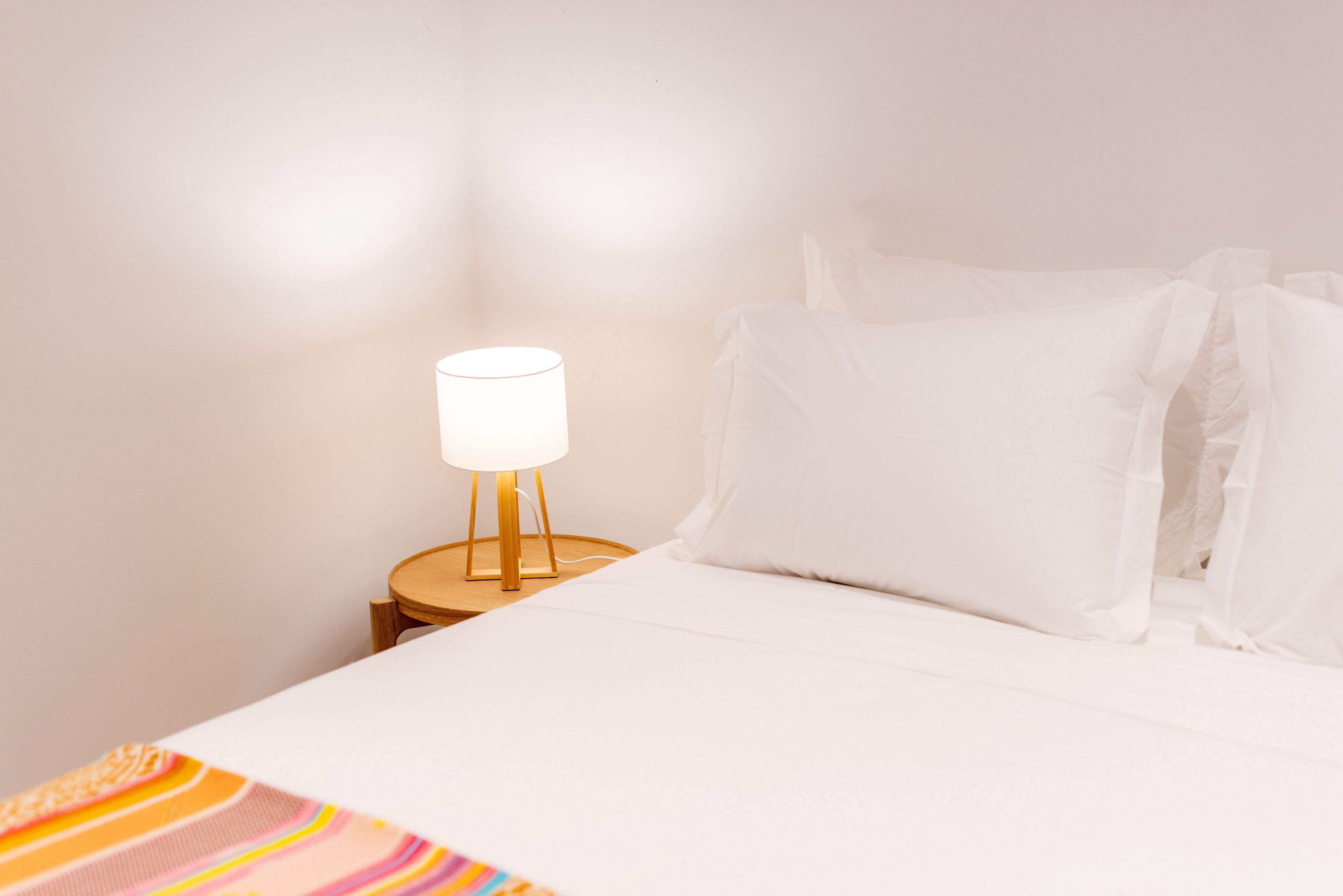 Ein ordentlich bezogenes Bett mit weißen Laken und einer Streifenwolldecke, eine Nachttischlampe und ein Nachttisch aus Holz.