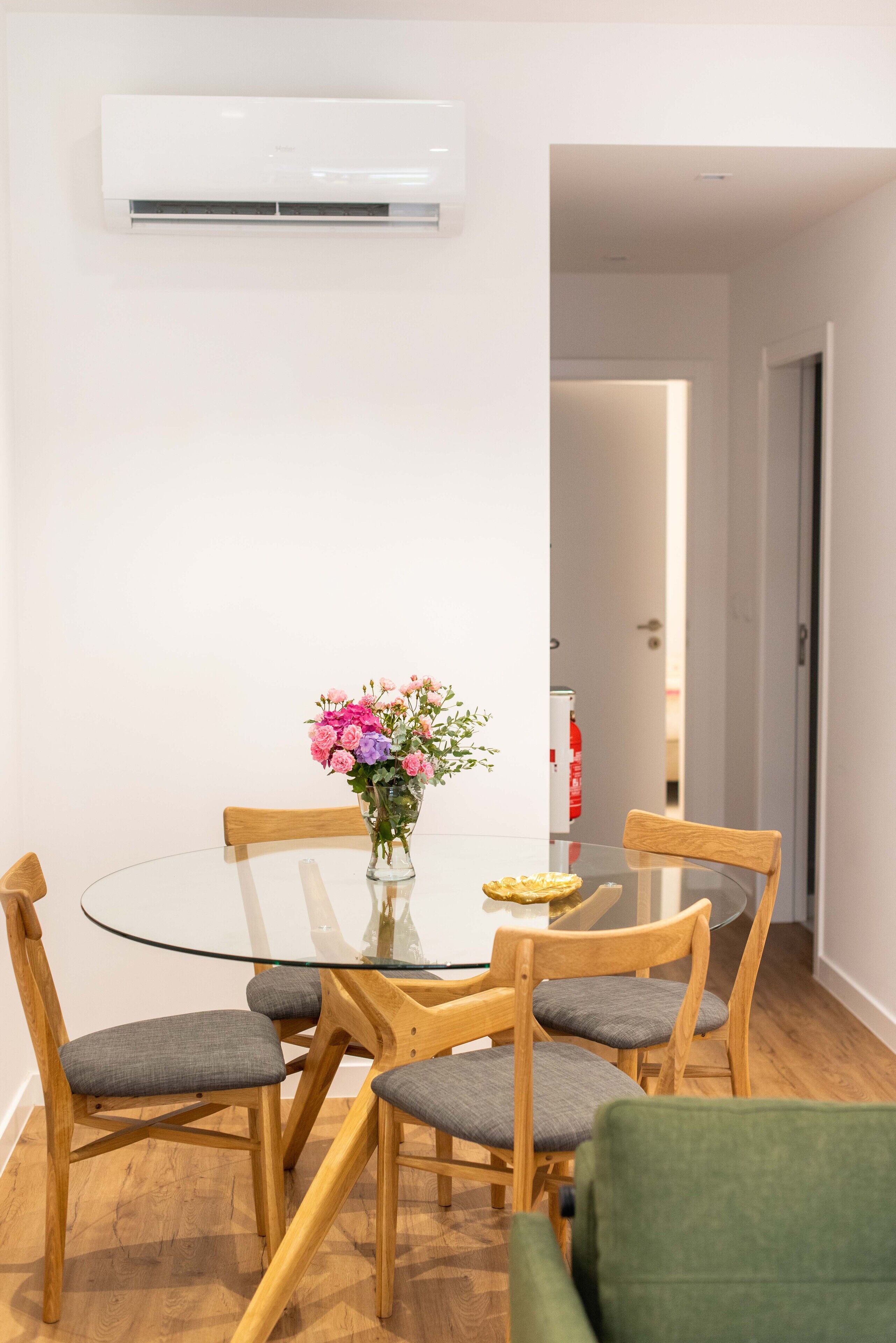 Spiseområde med et glasbord, træstole, en vase med blomster og en aircondition-enhed på væggen.
