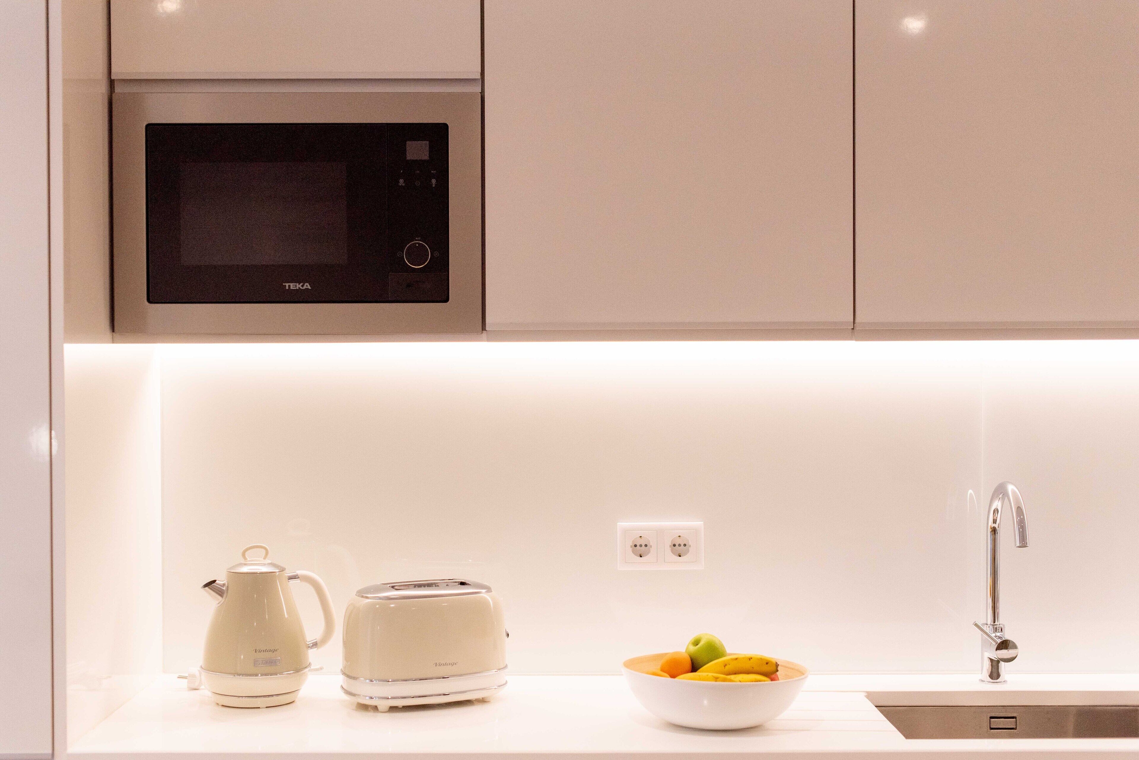 Une cuisine moderne équipée d’un four à micro-ondes, d’un grille-pain, d’une bouilloire et d’un saladier rempli de fruits posé sur le plan de travail.