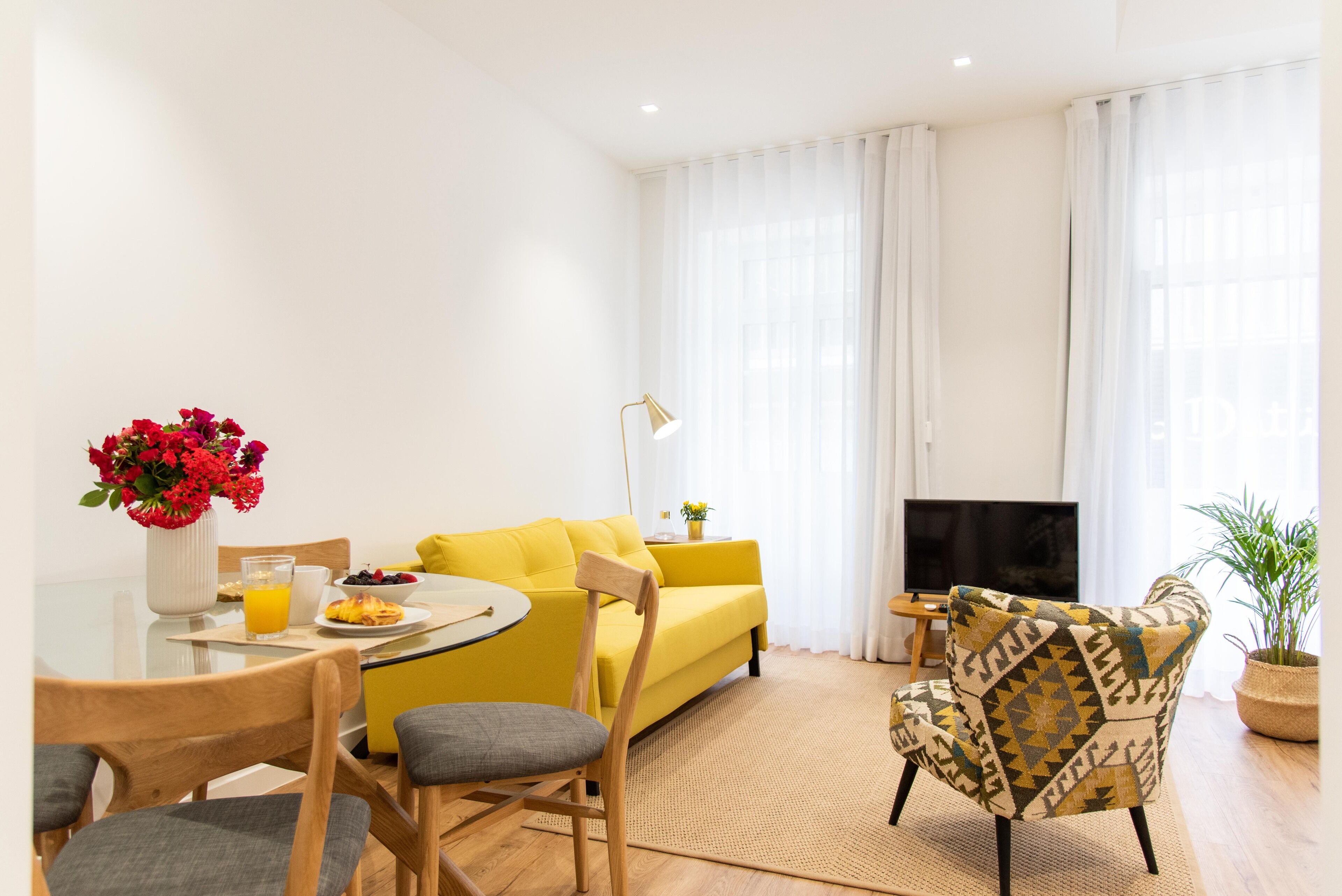 A bright living room with a yellow sofa, a dining table with chairs, a TV, and a potted plant.