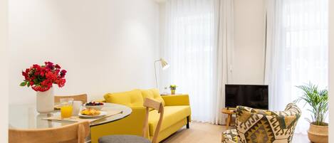 A bright living room with a yellow sofa, a dining table with chairs, a TV, and a potted plant.