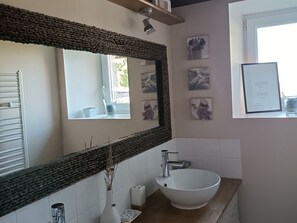 Une salle de bain avec un grand miroir, un lavabo blanc, une douche et une fenêtre ornée d’une œuvre d’art au mur.