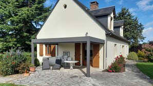 Ein modernes Haus mit einer Steinterrasse, einer überdachten Sitzgelegenheit im Freien und einem Garten mit verschiedenen Pflanzen und Blumen.