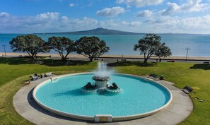 A circular pool with a central fountain, surrounded by a paved walkway, with trees and a view of the sea and a distant mountain in the background. - Sunshine Daydream w Edmund Cottage (Auckland)