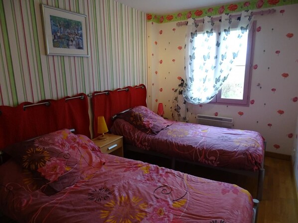 A bedroom with two beds, striped wallpaper, a framed picture on the wall, and a window with floral curtains. - Charming Cottage with Garden, Bikes & Fireplace Near Le Mans (Congé-sur-Orne)