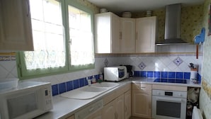 Une cuisine de taille modeste, équipée d’appareils électroménagers de base, une fenêtre ornée de rideaux en dentelle et un mur recouvert de carreaux.