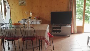 Un coin repas avec une table dressée pour quatre personnes, des chaises, un téléviseur posé sur un meuble et une fenêtre donnant sur un paysage verdoyant.