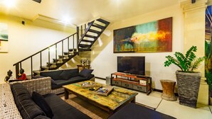 Un salon moderne avec un escalier, des canapés noirs, une table basse en bois, un téléviseur à écran plat et un grand tableau abstrait.