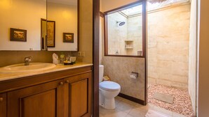 A bathroom with a wooden vanity, a large mirror, a toilet, and a shower area with stone walls. - Villa Arjuna (Seminyak)