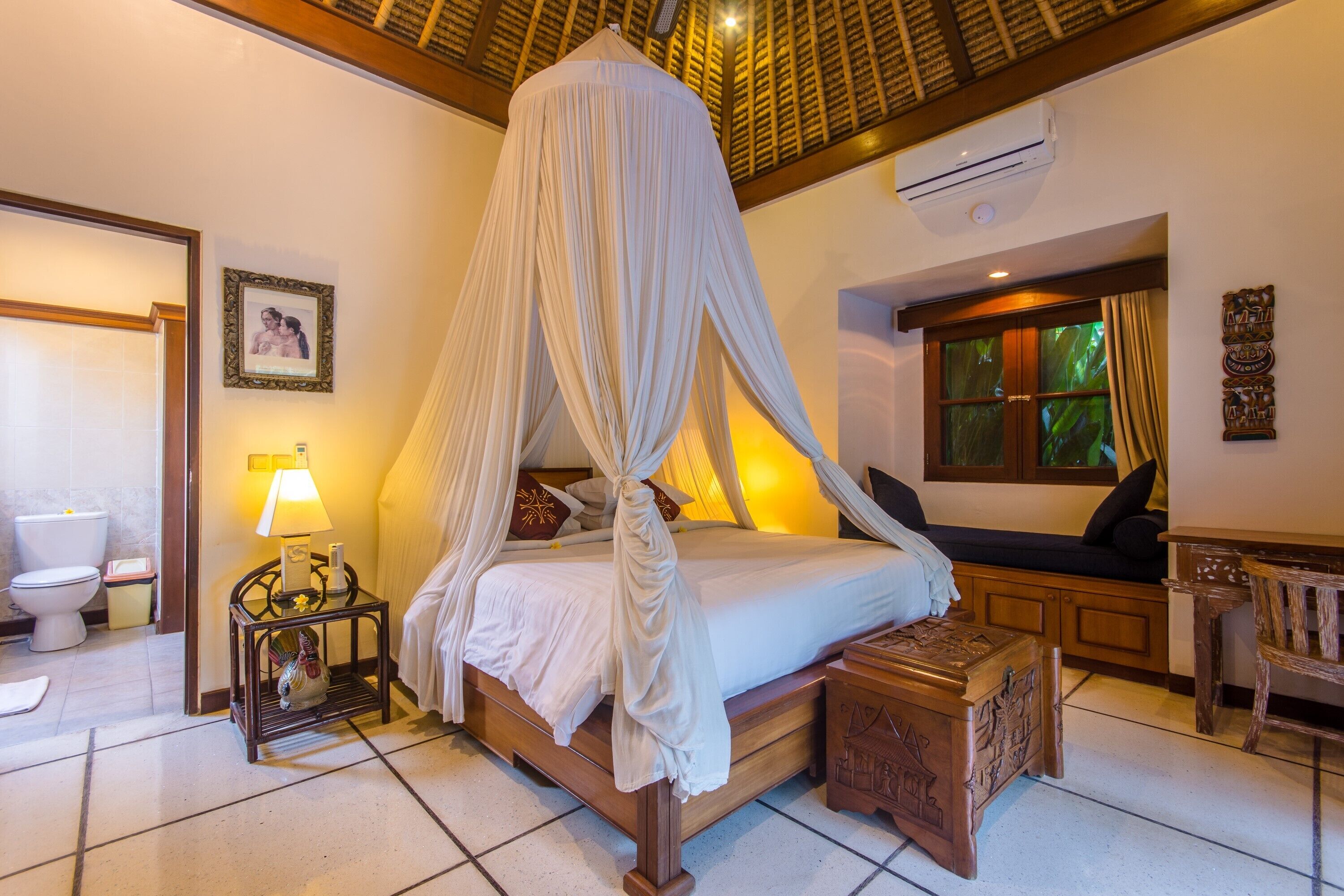 A bedroom with a four-poster bed, a bedside table with a lamp, a wooden chest, and a window with curtains.