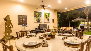 A dining area with a round table set for six, surrounded by wooden chairs. There are two paintings on the wall, a ceiling fan above, and a view of the outdoors with palm trees. - Villa Arjuna (Seminyak)