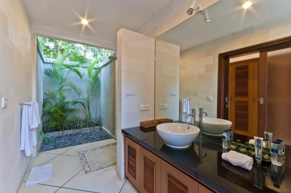 A modern bathroom with a large mirror, a white sink, and a shower area with a view of greenery.