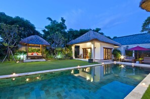 A poolside area with a clear pool, a thatched-roof structure, and a well-lit building with a tiled roof. - Villa Darma (Seminyak)