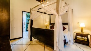 A four-poster bed with white drapes, a bedside table with a lamp, and a view of a garden through an open door. - Villa Kebun (Seminyak)