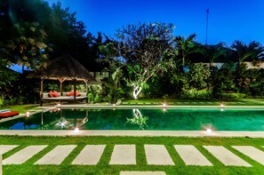 Une piscine agrémentée d’un gazebo à toit de chaume et de chaises longues, le tout entouré d’une végétation luxuriante et surplombé par un ciel nocturne dégagé.