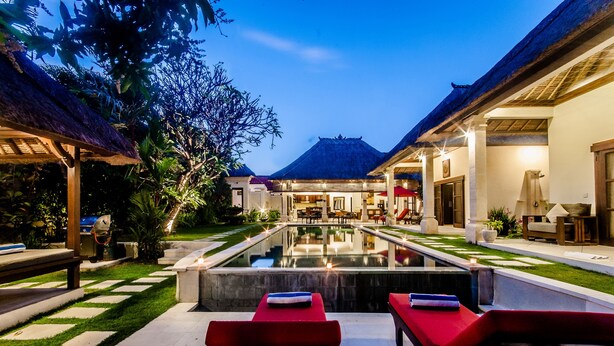 A pool area with lounge chairs, a thatched-roof structure, and a well-lit building in the background. - Villa Kebun (Seminyak)