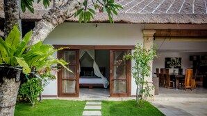 A house with a thatched roof, a visible bedroom with a bed and curtains, a dining area with chairs and a table, and a well-maintained lawn with stepping stones. - Villa Jaclan (Seminyak)